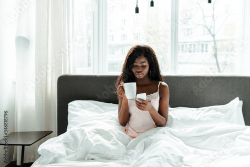 African american woman holding cup of tea and smartphone on bed in bedroom