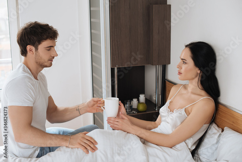 boyfriend giving cup of hot drink to ill girlfriend in bed during self isolation