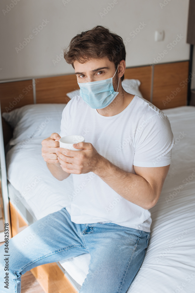 tired ill man in medical mask holding cup with hot drink on bed during self isolation tired ill man in medical mask holding cup with hot drink on bed during self isolation