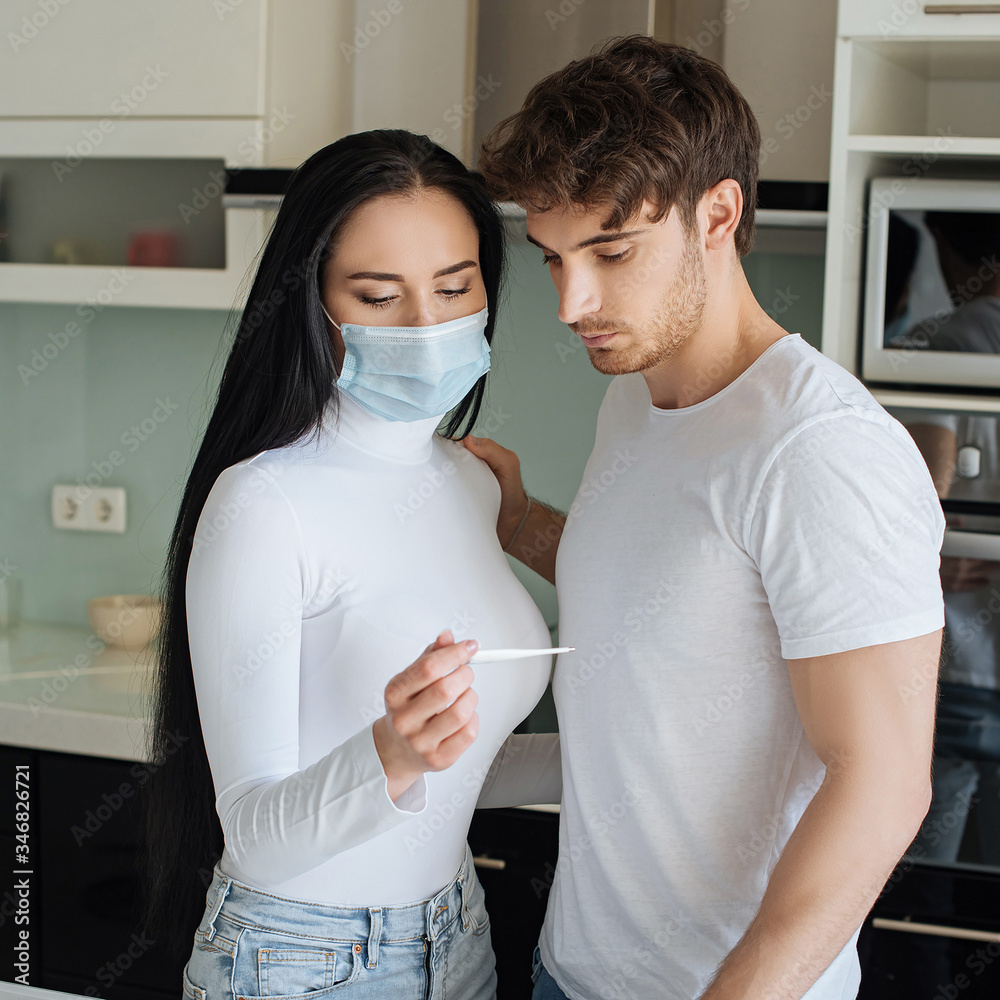 boyfriend and ill girl in medical mask looking at thermometer during self isolation