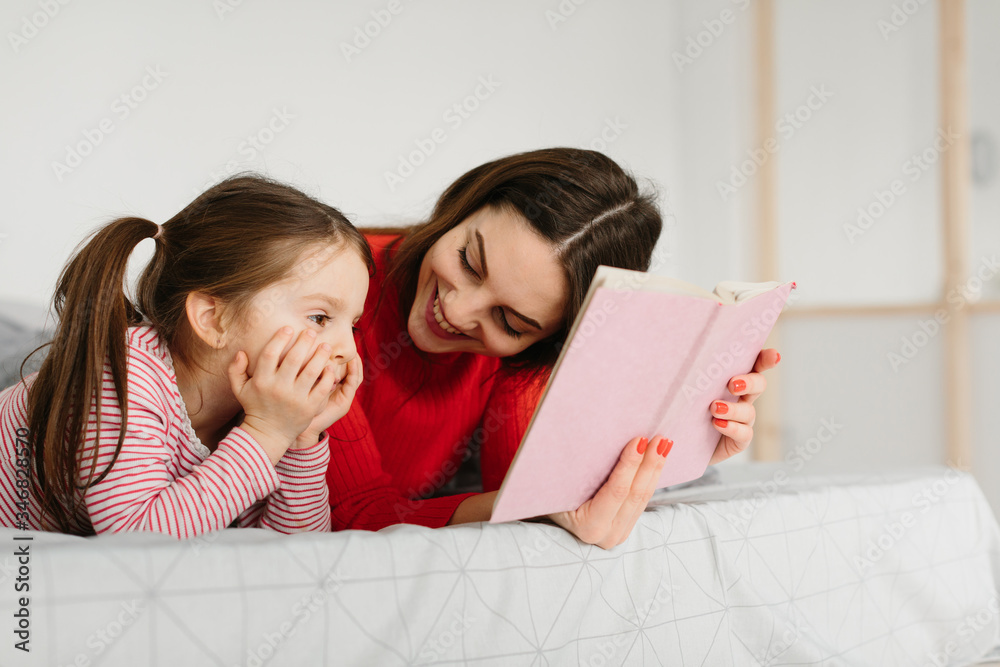 Happy family mother and child daughter reading holding book lying in bed, smiling mom baby sitter telling funny fairy tale to cute preschool kid girl having fun laughing together with bedtime stories