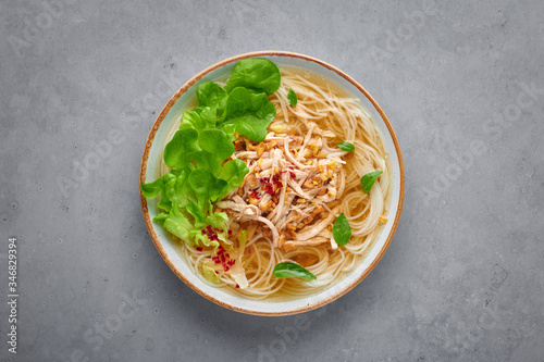 Guay Tiew Gai Cheek or Thai Chicken Noodle Soup in white bowl on gray concrete backdrop. Guay Tiew Gai is Thailand cuisine soup with rice noodles, chicken meat, sauces, greens. Thai food. Top view photo