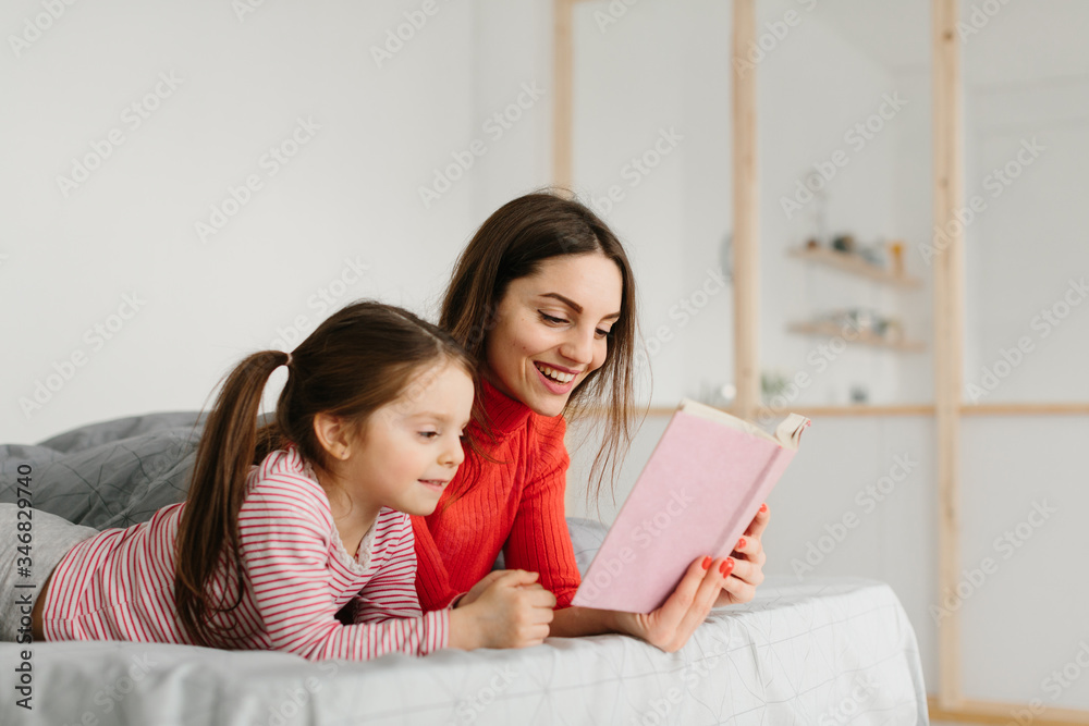 Happy family mother and child daughter reading holding book lying in bed, smiling mom baby sitter telling funny fairy tale to cute preschool kid girl having fun laughing together with bedtime stories Happy family mother and child daughter reading holding book lying in bed, smiling mom baby sitter telling funny fairy tale to cute preschool kid girl having fun laughing together with bedtime stories