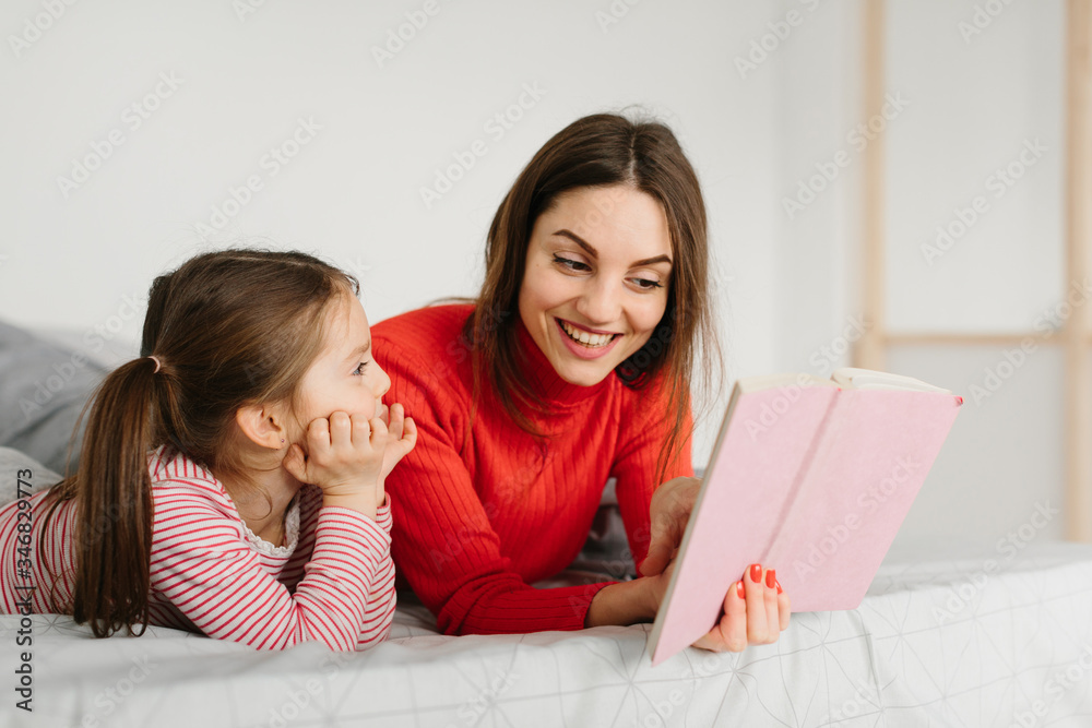 Happy family mother and child daughter reading holding book lying in bed, smiling mom baby sitter telling funny fairy tale to cute preschool kid girl having fun laughing together with bedtime stories Happy family mother and child daughter reading holding book lying in bed, smiling mom baby sitter telling funny fairy tale to cute preschool kid girl having fun laughing together with bedtime stories