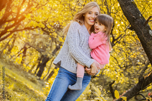 Happy little girl and her beautiful mother hugging each other and laughing while standing in autumn park. Young mother holding her daughter in hands while having fun together outdoors. Family love
