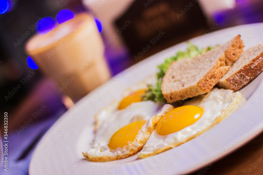 Closeup of fried eggs with toasted bread on white dish and glass with coffee on wooden table at modern restaurant. Traditional american breakfast in dark cafe Closeup of fried eggs with toasted bread on white dish and glass with coffee on wooden table at modern restaurant. Traditional american breakfast in dark cafe