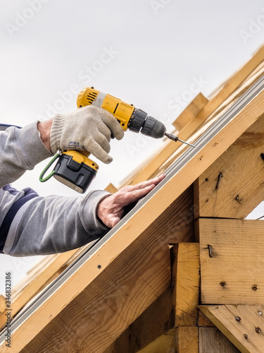 Men's hands in work gloves with a yellow screwdriver screw the roofing sheet to the roof of a country house. Cordless drill. The use of electrical engineering and technology.