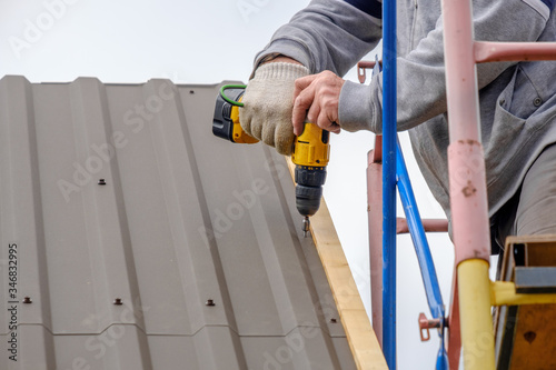 Men's hands in work gloves with a yellow screwdriver screw the roofing sheet to the roof of a country house. Cordless drill. The use of electrical engineering and technology.