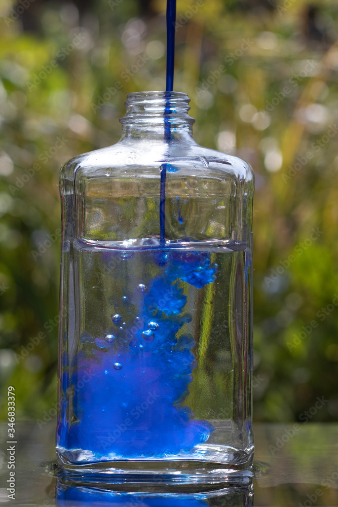 Blue food colouring being poured into water in a rectangle shaped bottle. 