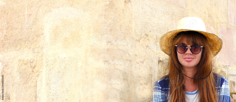 Close up of beautiful tourist woman with long red hair, sunglasses and a hat. Pretty girl with hat in Spain on the wall background. Panoramic banner view of traveler girl