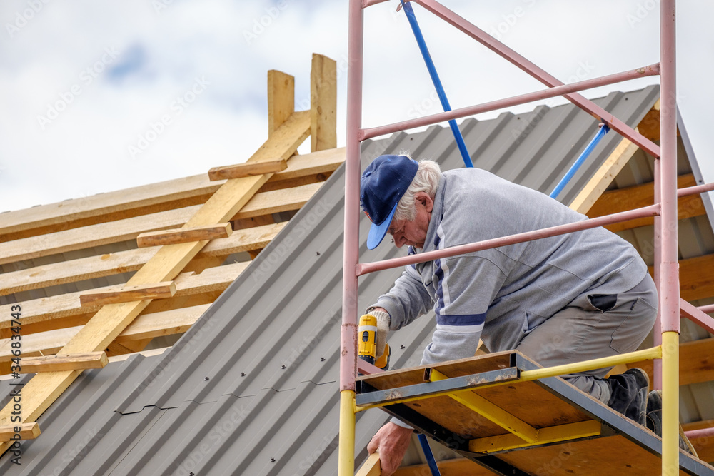 Senior gray-haired Construction man using a screwdriver, fastens a roofing sheet to wooden rafters on the roof of a country house under construction. The physical activity of the elderly. Senior gray-haired Construction man using a screwdriver, fastens a roofing sheet to wooden rafters on the roof of a country house under construction. The physical activity of the elderly.