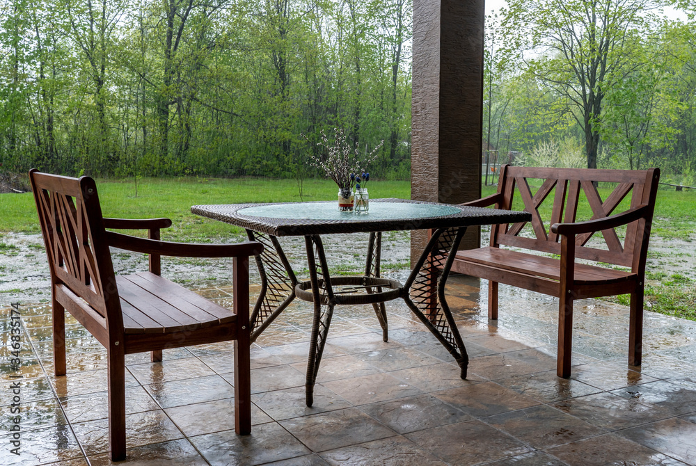 Two wooden benches and a table stand on the outdoor verandah during heavy rain