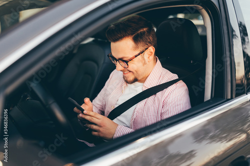 Side view through open window of a driver sitting in a car and looking in smartphone. A smiling happy young man with beard and stylish glasses © Kate