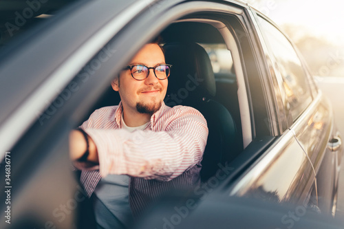 A young happy stylish smiling man in glasses driving a car and looking out open window