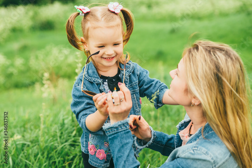 Blonde mother and little daughter walk in nature, in the meadow. Mother and daughter dressed in denim jackets, mother took off her glasses and gives their daughter to dress. photo