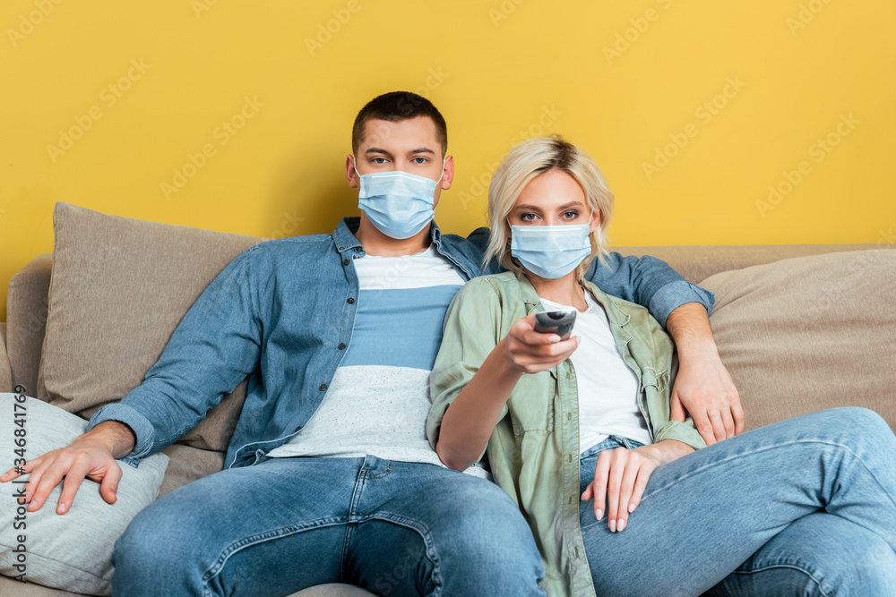 young couple in medical masks watching tv on sofa near yellow wall