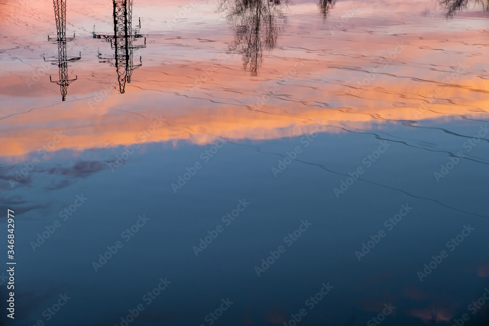 Reflection of a power transmission pylon and orange clouds in water.