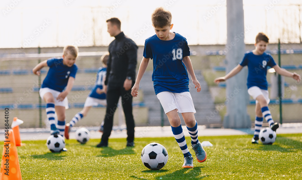 Fototapeta premium Portrait of school boys training soccer with young coach on football field. Boys running with football balls between colorful soccer markers and cones. Practice sessnion for youth soccer team.