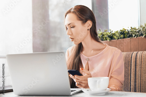 Focused woman is sitting at table in front of panoramic window, working at laptop in cafe, well-groomed beautiful beauty view, wears delicate pink shirt. A freelancer works remotely at computer phone