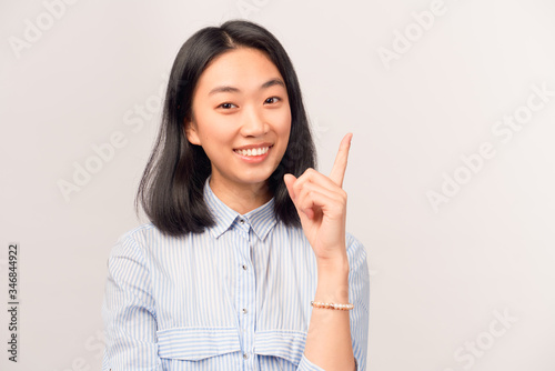Close-up of young brunette woman pretty pointing her finger up has brilliant idea, sign gesture. Portrait of a girl looking at camera isolated on white wall in a Studio wearing striped shirt, Eureka