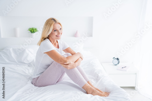 Profile side view portrait of her she nice attractive lovely cheerful cheery aged woman sitting on bed living healthy life lifestyle young soul in modern light white interior room flat apartment