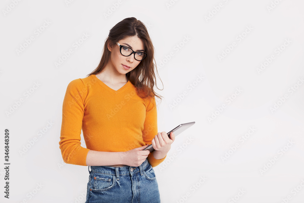 A young girl in glasses and a yellow sweater is holding a tablet and smiling. Portrait of a girl on a light background with space for text A young girl in glasses and a yellow sweater is holding a tablet and smiling. Portrait of a girl on a light background with space for text