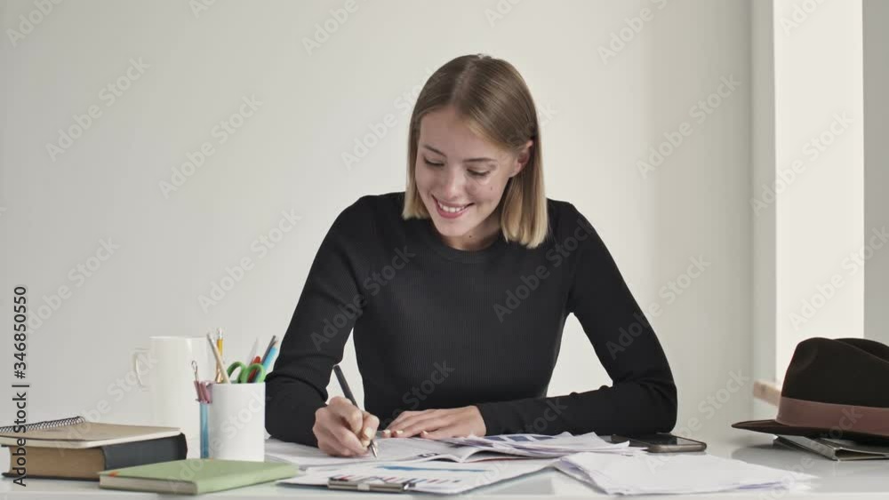 A beautiful young blonde woman is writing something while comes up with a good idea sitting at the table indoors in a white office A beautiful young blonde woman is writing something while comes up with a good idea sitting at the table indoors in a white office