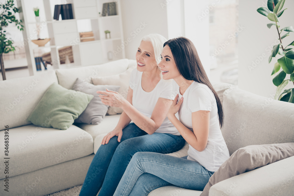 Portrait of two nice attractive lovely cheerful cheery women sitting on divan watching interesting tv show cinema film spending free spare time day in white light interior house flat apartment