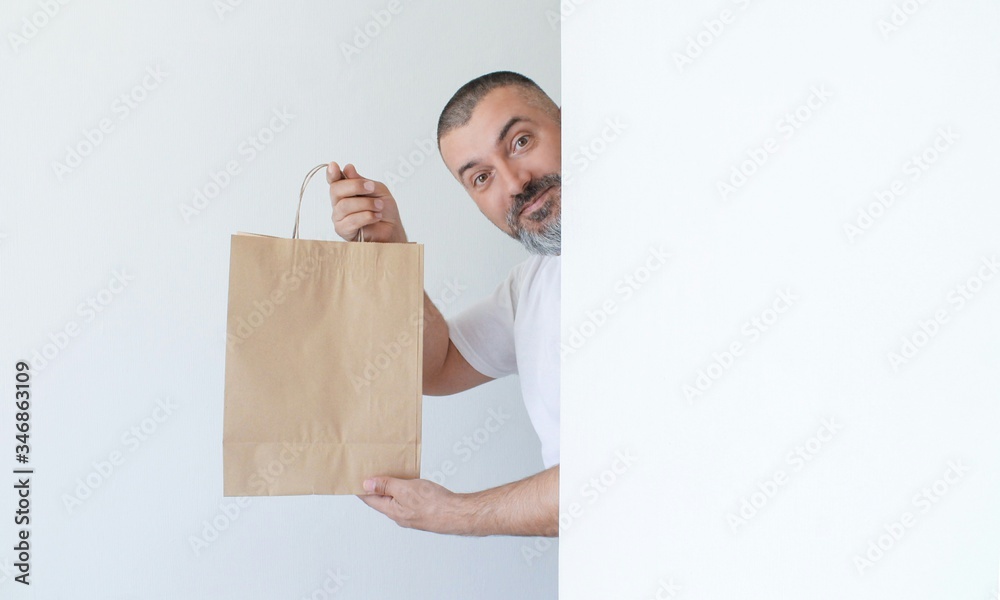 Handsome smiling man with beard peeking behind a white wall and holding paper bag in hands isolated on white background Handsome smiling man with beard peeking behind a white wall and holding paper bag in hands isolated on white background