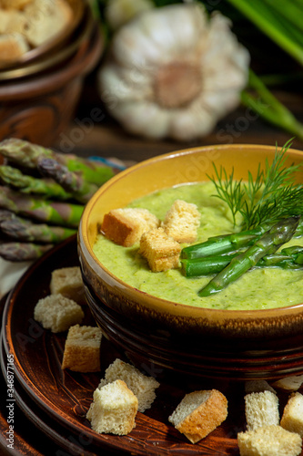 Purred creamy asparagus soup in glass bowl on black plate against raw fresh asparagus and greenery photo