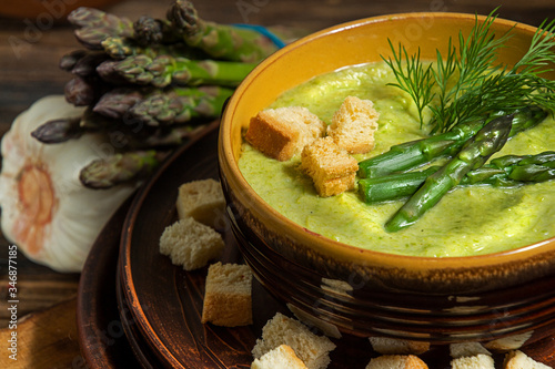 Purred creamy asparagus soup in glass bowl on black plate against raw fresh asparagus and greenery photo