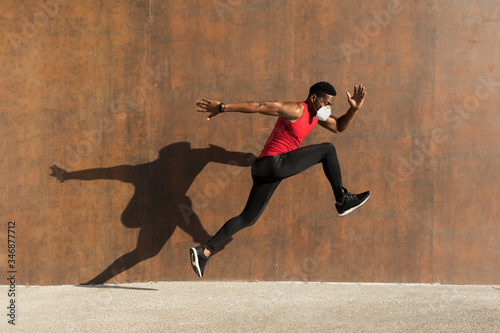 Powerful athlete training with n95 face mask for protecting against Covid-19. Young black man running and jumping casting shadow on a wall.
