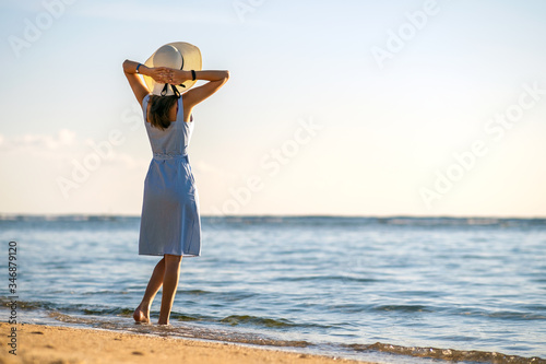 Young woman in straw hat and a dress standing alone on empty sand beach at sea shore. Lonely tourist girl looking at horizon over calm ocean surface on vacation trip.