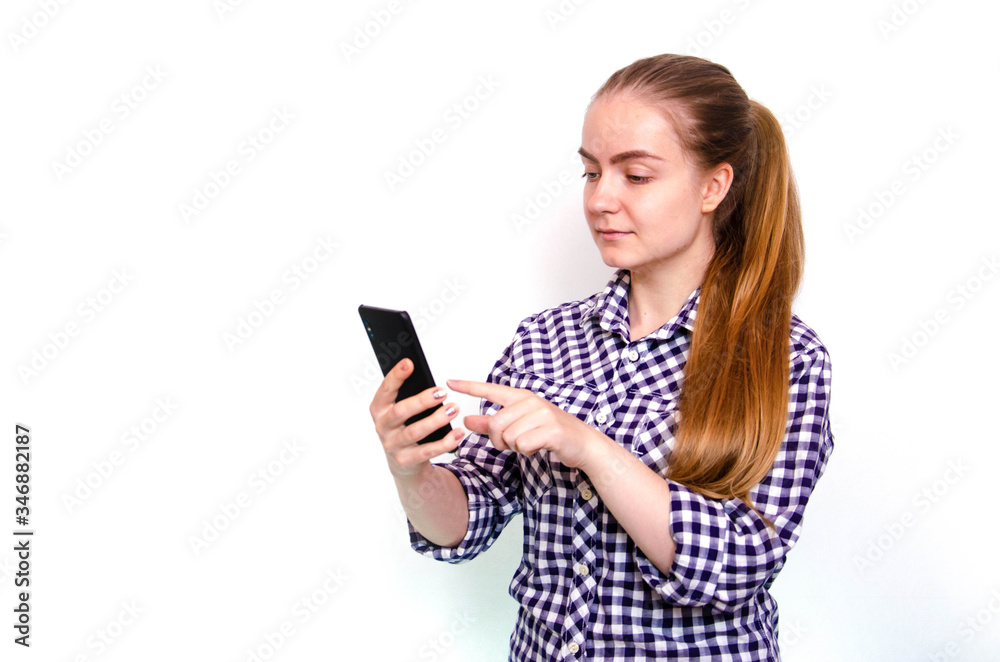 A young girl in a plaid shirt with long hair stands on a white background and holds a smartphone in his hand. A girl looks, is surprised, scared, rejoices looking at a smartphone A young girl in a plaid shirt with long hair stands on a white background and holds a smartphone in his hand. A girl looks, is surprised, scared, rejoices looking at a smartphone