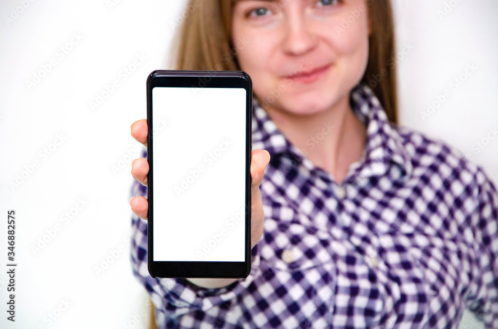 A young girl in a plaid shirt with long hair stands on a white background and holds a smartphone in his hand. Shows smartphone screen, white screen. A girl shows something on a  smartphone.