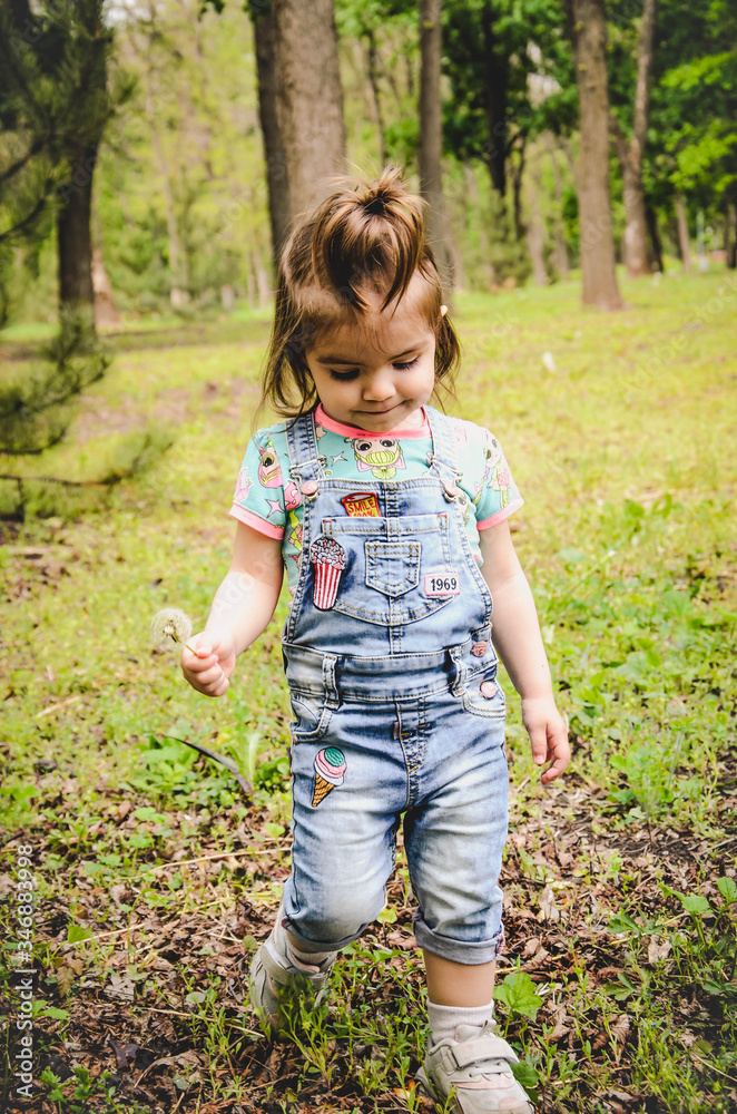 May 2020. Kiev Ukraine. A little girl plays outside in a park, collects plants on a sunny day in denim overalls and a T-shirt with a print LOL May 2020. Kiev Ukraine. A little girl plays outside in a park, collects plants on a sunny day in denim overalls and a T-shirt with a print LOL