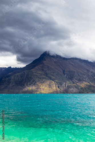 Mountain peaks up to clouds over turquoise water. Rainy day at Lake Wakatipu, New Zealand