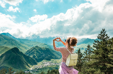Young female making hands in heart shape sign on the peak of mountains. View in the top of mountain on a hot summer day. Lifestyle concept. Back view. mountain landscape. Freedom concept.