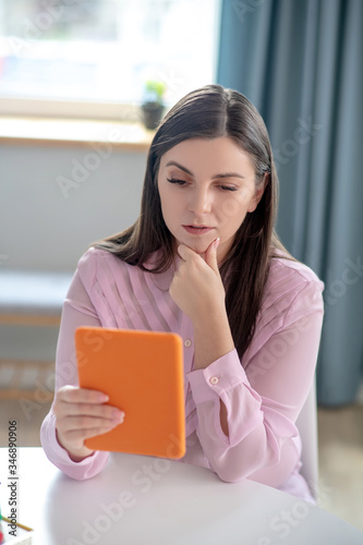 Pretty young woman in a pink blouse sitting at the table and looking thoughtful