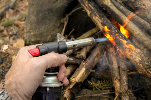 Man with a butane can burner kindles a fire in a bonfire in forest