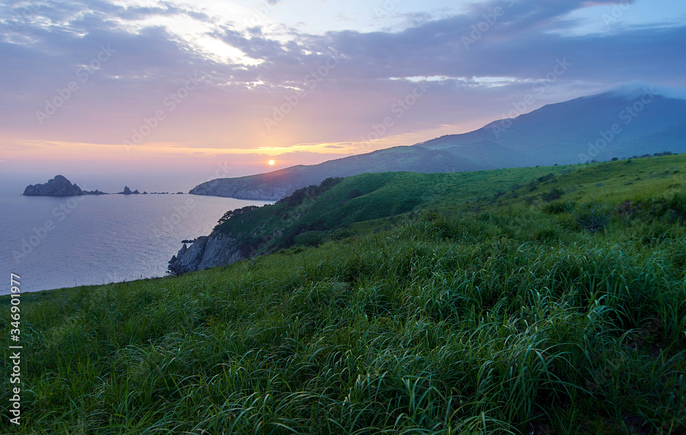 Green hills and steep rocky shores by the ocean. View of the rugged islands and peninsulas at sunset. In the foreground is green grass.