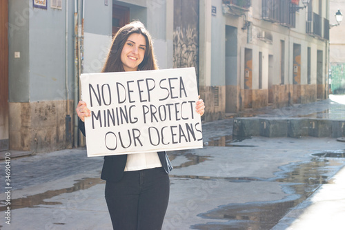 attractive  young woman activist hold up protesting sign saying "No deep sea mining protect our oceans" in the city, dark air. Place for your text in copy space.