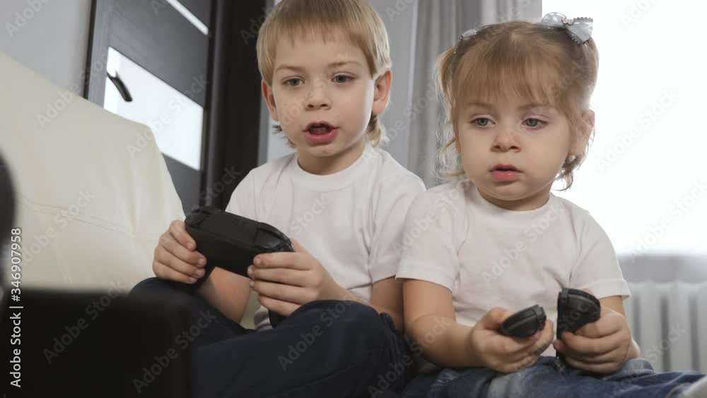 Blonde-haired children a girl and a boy play a game console. Happy children play sitting on a white sofa at home. Blonde-haired children a girl and a boy play a game console. Happy children play sitting on a white sofa at home.