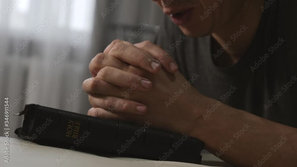 A man prays with his hands crossed over a Bible for God's blessing in a dark room.