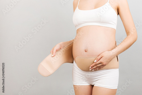 Close up of young pregnant woman in underwear dressing bandage on her belly at gray background with copy space. Orthopedic abdominal support belt concept