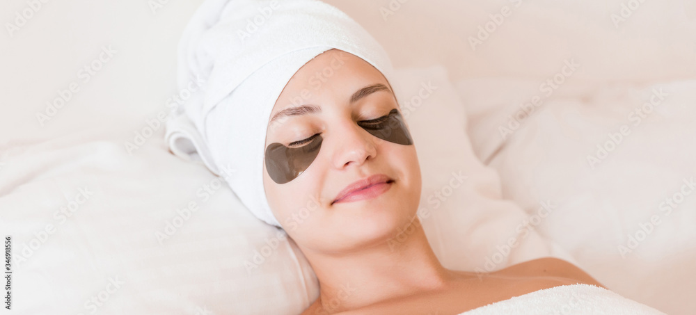 Young woman with closed eyes and black eye patches is lying and relaxing in the bed after having a bath wrapped in towel. Beauty treatment and skincare concept