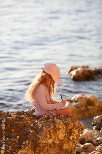 Fototapeta Naklejka Na Ścianę i Meble -  Happy pretty girl walks along the sea coast against the background of the sea, from behind a beautiful landscape. The child draws in a notebook