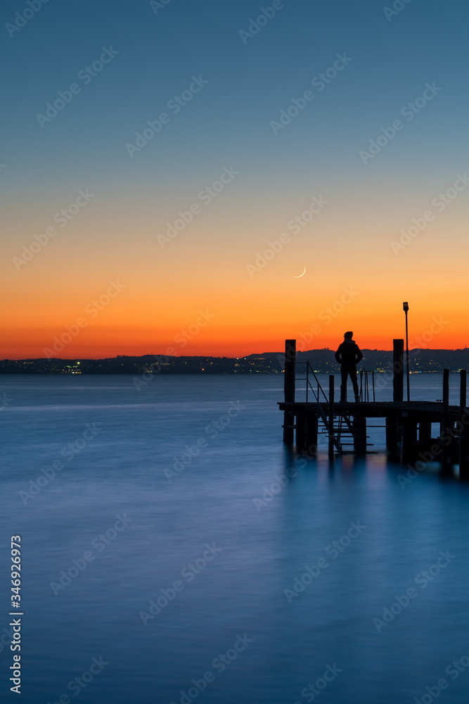 A man silhouette standing on wooden pier lonely at the sea with beautiful sunset. lsunset seascape at a wooden jetty.