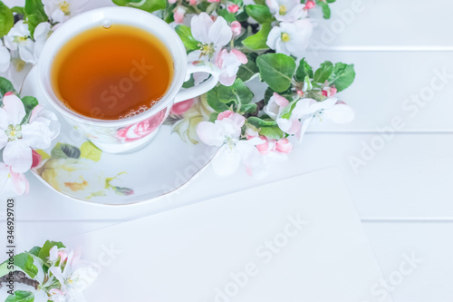 a cup of tea, a piece of paper and white flowers on a white background. a cup of tea and branches of a blooming apple tree close-up. festive background with a cup of tea and white flowers.