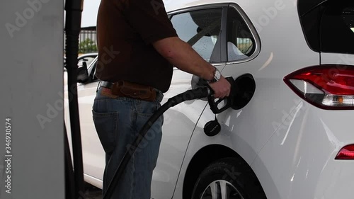 man filling the tank a white car. wearing blue jeans and a brown polo